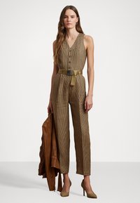 Combinaison à carreaux bruns avec un décolleté en V profond, dotée de boutons, d'une taille ceinturée et de jambes droites. Associée à une veste en cuir marron.