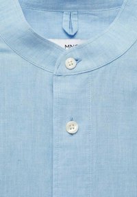 Lichtblauwe linnen shirt met een staande kraag, voorzien van een knoopsluiting aan de voorkant met twee witte knopen en een klein haakje bij de halslijn.