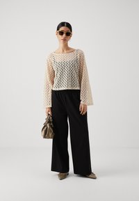 Haut à manches longues beige à motif en treillis, pantalon noir à jambes larges et ballerines beiges à bout pointu. Tenant un petit sac métallique, portant des lunettes de soleil.
