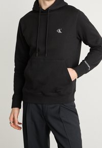 Black cotton hoodie featuring a kangaroo pocket, drawstrings at the neck, and a white logo on the chest and sleeve. Textured fabric.