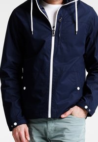 Marinblå zip-jacka i lätt tyg, med en vit dragkedja, snoddar, två sidofickor och knappstängningar vid ärmarna.