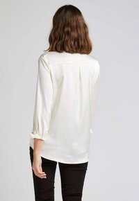 Blouse blanche à manches longues avec une texture lisse, dotée d'un empiècement au dos, de poignets retroussés et d'un subtil motif à pois. Pantalon noir visible en dessous.