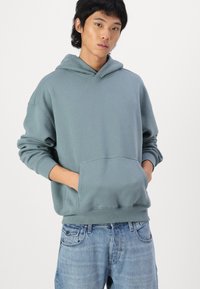 Felpa con cappuccio in morbido tessuto blu-verde, con tasca frontale e polsini a costine, abbinata a jeans in denim blu chiaro.