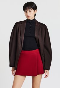Giovane con capelli corti e scuri indossa una camicia nera a bottoni, un blazer oversize marrone scuro e una gonna corta rossa brillante, su uno sfondo neutro.