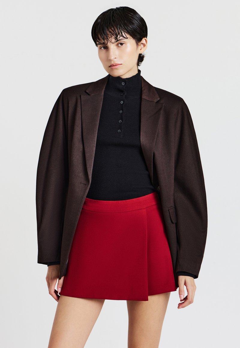 Giovane con capelli corti e scuri indossa una camicia nera a bottoni, un blazer oversize marrone scuro e una gonna corta rossa brillante, su uno sfondo neutro.
