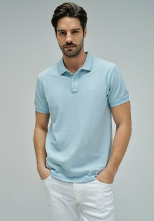Hombre con cabello corto y oscuro y barba, vestido con una camiseta polo azul claro y pantalones blancos, de pie con las manos en los bolsillos frente a un fondo gris.