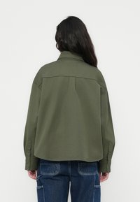 Κοντό, χακί πράσινο μπουφάν από μαλακό ύφασμα. Διαθέτει αιχμηρό γιακά, μανσέτες με κουμπιά και ίσια γραμμή στο πίσω μέρος.