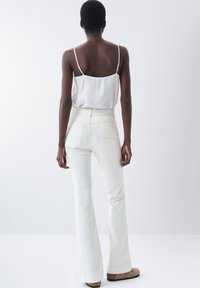 Jeans blancos acampanados con bolsillos traseros, de cintura alta y textura suave, combinados con una blusa ligera sin mangas. Modelo de espaldas, con tirantes sutiles visibles.