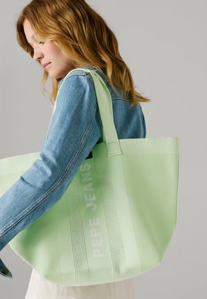 Nuori nainen farkkutakissa kantamassa suurta vaaleanvihreää Pepe Jeans -olkalaukkua yksiväristä taustaa vasten.