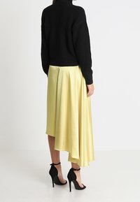 Femme debout, portant un pull noir, une jupe midi asymétrique jaune et des sandales à talons hauts noires avec brides à la cheville.