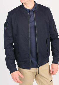 Homme portant une veste bomber bleu marine zippée par-dessus un polo bleu foncé et un pantalon beige, se tenant avec les mains détendues le long du corps.