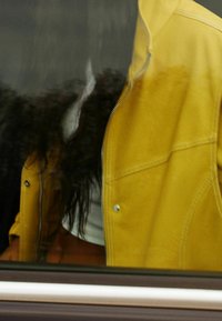 Osoba w żółtej kurtce i białej bluzce, widoczna przez okno z odbiciem na szybie.