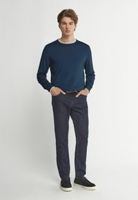 Mörkblå långärmad tröja med vit rundhals, tillsammans med mörka denimjeans och svarta sneakers med vit sula. Stående pose.