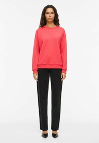 Sweat-shirt rouge à col rond, épaules tombantes et poignets côtelés, associé à un pantalon droit noir taille haute et des chaussures noires à bouts pointus.