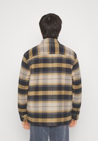 Károvaná košile v béžových, námořnických a žlutých odstínech; volný střih s límcem na knoflíky; látka vypadá texturovaně a teple.