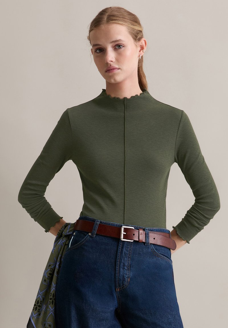 Langsleeve, hoge hals groene geribbelde top met gegolfde randen, gecombineerd met donkere spijkerbroek en een bruine riem. Stevige textuur stof.