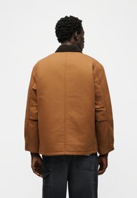 Άνδρας με καφέ μπουφάν με σκούρο γιακά και μαύρο παντελόνι, όρθιος με την πλάτη προς το μέρος μας, μπροστά σε απλό λευκό φόντο.