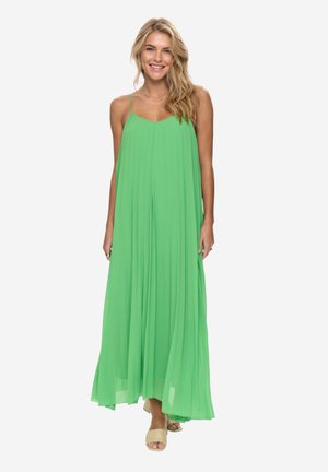 Blondynka uśmiechająca się, ubrana w luźną, jasnooliwkową, długą sukienkę bez rękawów na cienkich ramiączkach oraz beżowe sandały na odkryte palce, stojąca na białym tle.