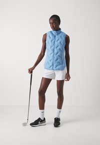 Svijetloplavi bezrukavnik s punjenjem i kocinim dizajnom, zatvaranje na patentni zatvarač i visokim ovratnikom, u kombinaciji s bijelim šortsevima i crnim tenisicama.