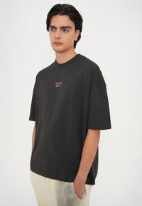 Camiseta de algodón negra con mangas cortas, que presenta un pequeño logo púrpura de Reebok en el pecho. Diseño de ajuste relajado y cuello redondo.