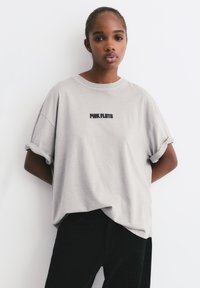 Młoda osoba z warkoczami, ubrana w jasnoszary t-shirt z logo Pink Floyd i czarne spodnie, stoi na białym tle.