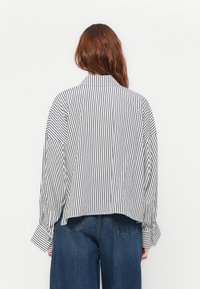 Frau mit langen rötlichen Haaren, die ein lockeres, langärmliges weißes Hemd mit schwarzen vertikalen Streifen und blaue Jeans trägt, mit dem Rücken zugewandt.