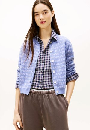 Junge Frau trägt eine blaue Tommy Jeans Strickjacke über einem hochgekrempelten karierten Hemd und lockeren braunen Hosen mit den Händen in den Taschen.