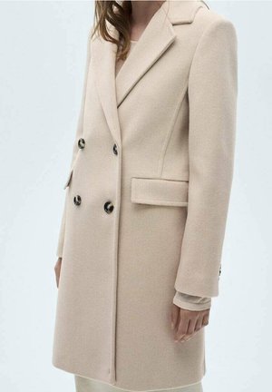 Mujer con un abrigo de lana beige de doble botonadura con botones negros y bolsillos de solapa, de pie frente a un fondo claro y liso.