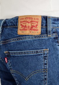 Blå denimjeans med en lærlabel med rød tekst. Labelen viser merkeinformasjon, pleieinformasjon og et registrert varemerke.