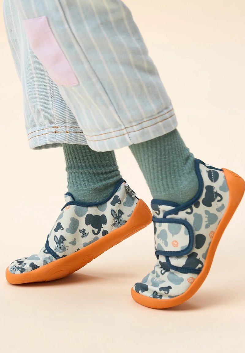 Enfant portant des chaussures bleu clair à motifs avec des semelles orange et des chaussettes vertes, debout sur une surface beige.
