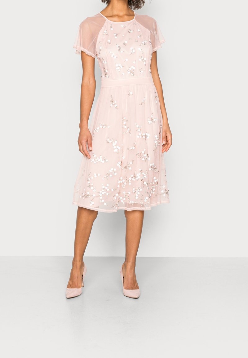Femme portant une robe rose pâle longueur genou avec des manches courtes transparentes et une broderie florale blanche, assortie à des talons roses.