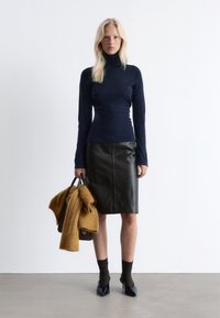 Marinefarbener Rollkragenpullover, schwarze, kroko-embossierte Leder-Bleistiftrock, braune Stricksocken und spitze Schuhe, in der Hand eine braune Tasche und einen gelben Pullover.