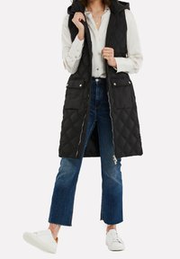 Femme portant une longue veste matelassée noire à capuche par-dessus un chemisier blanc, un jean évasé court bleu, et des baskets blanches, debout avec un pied levé.