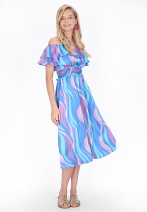 Donna sorridente con lunghi capelli biondi che indossa un vestito midi blu e rosa a fantasia con spalle scoperte e orecchini circolari rosa, in piedi su uno sfondo bianco.