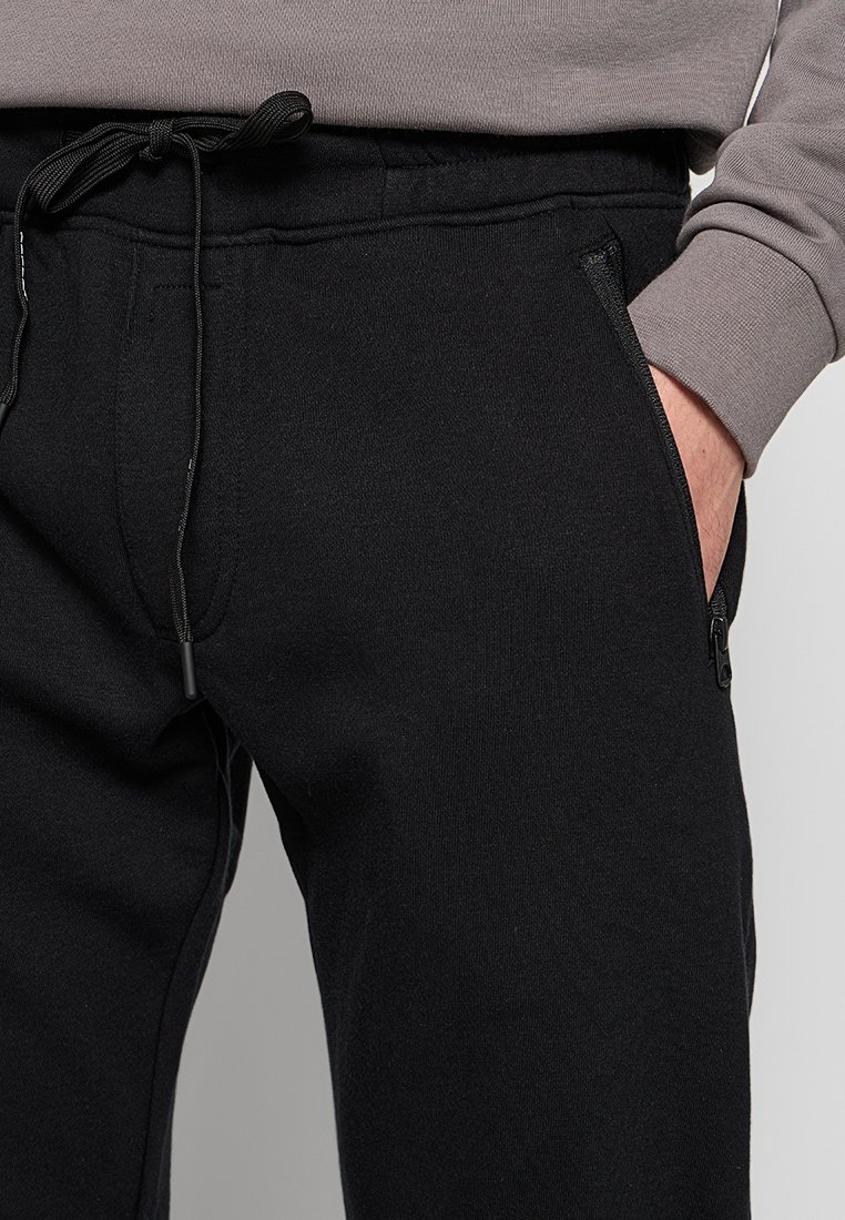 Pantaloni della tuta neri con vita regolabile, dotati di tasche laterali e una texture liscia. Il tessuto è morbido con una sensazione leggermente elastica.