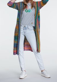 Cardigan tricot multicolore à manches longues et poches, porté sur un t-shirt gris avec texte, associé à un jean bleu clair et des baskets blanches.