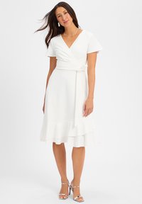Witte wikkeljurk met korte mouwen, een strik in de taille en een gelaagde ruffled zoom. Het model draagt zilveren hakken en een ketting.