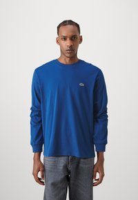 T-shirt en coton bleu à manches longues avec un col rond et des poignets côtelés, arborant un petit logo vert sur la poitrine gauche. Porté avec un jean gris.