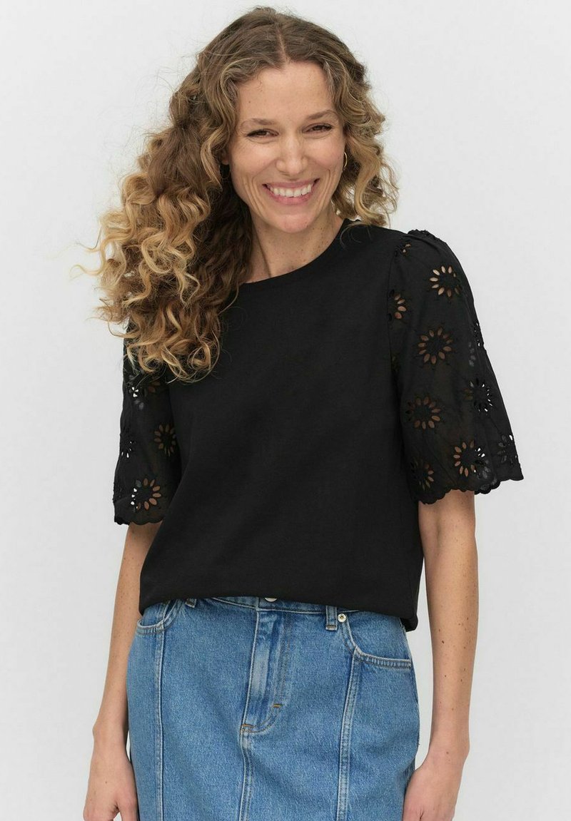 Czarna bluzka z haftowanymi, bufkowatymi rękawami z kwiatowymi wycięciami; zestawiona z dżinsową spódnicą. Prosty fason z teksturowanymi detalami.