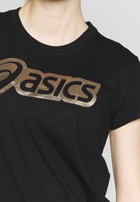 Camiseta negra de algodón con un logo "Asics" metálico en color dorado en el pecho. Mangas cortas y diseño de cuello redondo.