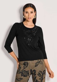 Schwarzes Langarmoberteil mit runder Halslinie, das mit einem erhabenen Blumendesign verziert ist, das mit Pailletten geschmückt ist. Das Modell kombiniert es mit gemusterten Hosen.