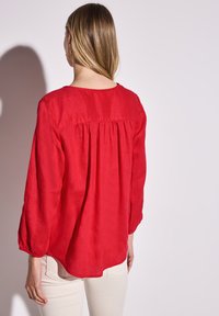 Blusa rossa a maniche lunghe realizzata in tessuto leggero, con scollo rotondo e dettaglio arricciato sul retro. Le maniche presentano polsini elastici.