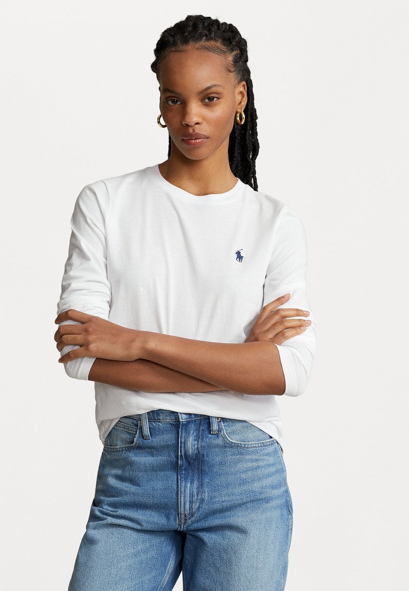 Langärmliges, weißes T-Shirt aus Baumwolle mit einem kleinen blauen Logo auf der linken Brust, kombiniert mit hochgeschnittenen blauen Jeans.