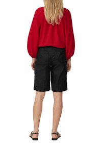 Rote strukturierte Bluse mit Puffärmeln, kombiniert mit schwarzen knielangen Shorts. Schwarze Sandalen runden das Outfit ab und zeigen einen lässigen Stil.