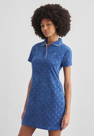 Femme avec une coiffure afro portant une robe polo bleu texturée à manches courtes avec un bord blanc et un petit logo Nike sur la poitrine.