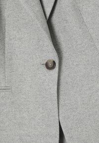 Manteau gris clair à motif chevrons avec un seul bouton marron foncé fermé et une poche passepoilée latérale visible.