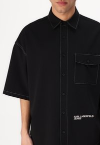 Juodas trumpomis rankovėmis marškinėliai su sagčių užsegimu, kontrastinga balta siūlė, krūtinės kišenė ir logotipo detalė priekyje.