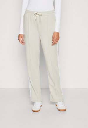 Personne portant un pantalon beige à cordon avec une bande blanche sur le côté, associé à des baskets blanches et un haut blanc à manches longues.