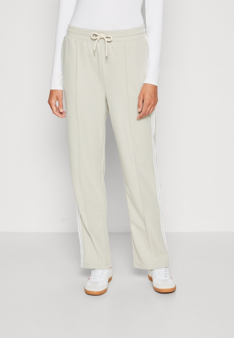 Personne portant un pantalon beige à cordon avec une bande blanche sur le côté, associé à des baskets blanches et un haut blanc à manches longues.