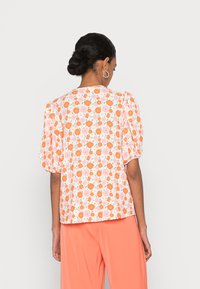 Blusa floral em laranja e rosa sobre tecido branco, com mangas bufantes e decote redondo. Combinada com calças largas em cor coral.